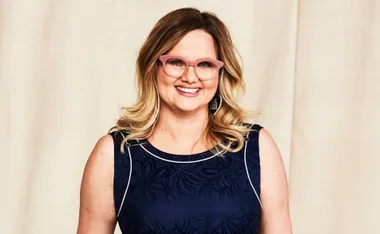 A woman wearing pink glasses and a navy dress smiles against a beige background.