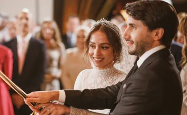 A bride wearing a lace dress and tiara smiles beside the groom in a suit, surrounded by guests at a wedding.