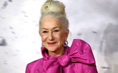 Woman with long white hair in an elegant updo, wearing a bright pink outfit with a large bow, smiling warmly.