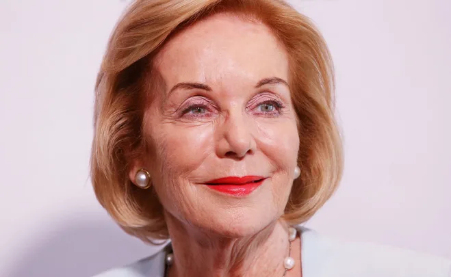 Elderly woman with blonde bob hairstyle, wearing pearl earrings and red lipstick, smiling against a neutral background.