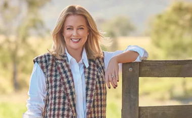 Woman smiling by a wooden fence in a rural setting, wearing a plaid vest and white shirt.
