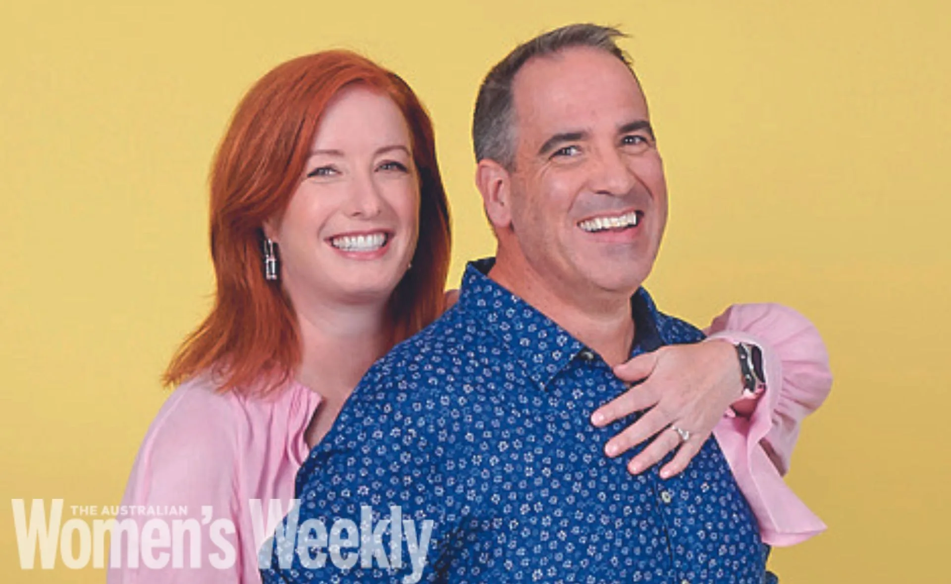 Smiling man in a blue patterned shirt with a woman in pink behind him on a yellow background.