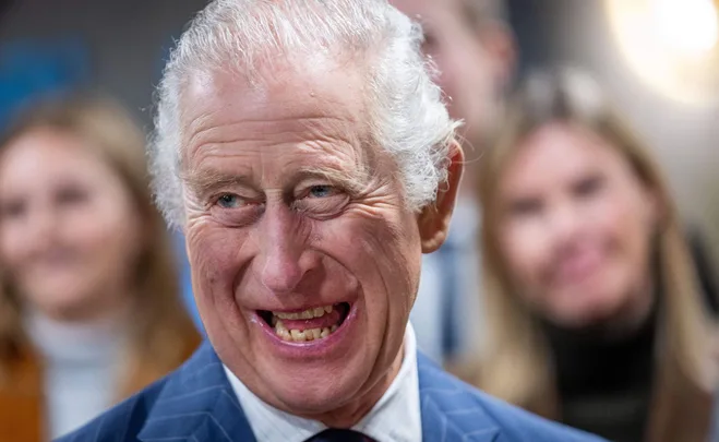 A man in a blue suit smiling broadly with a few people blurred in the background.