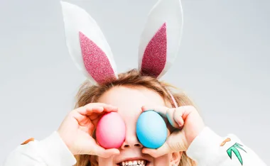 Child with pink bunny ears holding pink and blue Easter eggs over their eyes, smiling broadly in a playful Easter-themed setting.