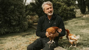A man sitting outdoors holding a chicken, with another chicken nearby, surrounded by greenery.