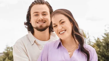 A couple smiling outdoors; the woman wears a purple shirt, the man a light shirt.