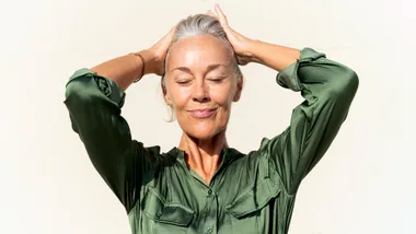 Woman in a green shirt with closed eyes and hands on head, smiling contently.
