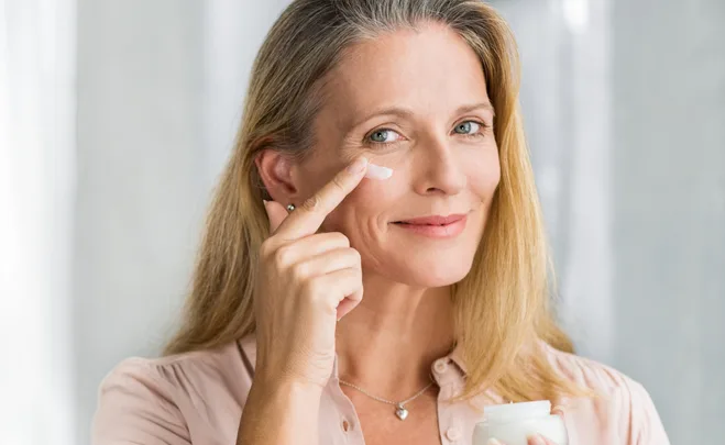 Woman applying cream under her eyes, smiling, with light background.