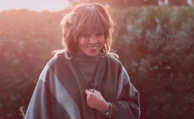 A woman stands smiling outdoors with a warm, sunlit background, wearing a cozy shawl.