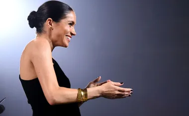 A woman in a black dress, side profile, gesturing with her hands, wearing gold bracelets and dark nail polish.