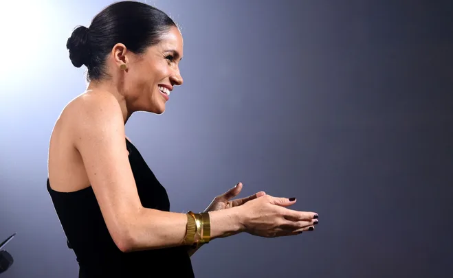 A woman in a black dress, side profile, gesturing with her hands, wearing gold bracelets and dark nail polish.