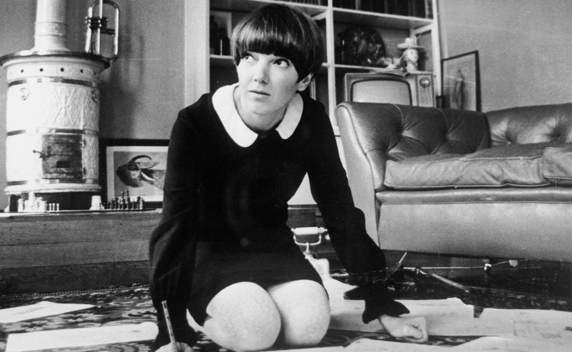 Designer Mary Quant kneeling on a carpet, sketching designs, in a room with vintage decor.