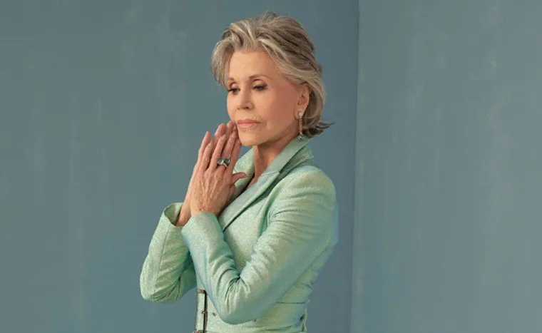 Elegant woman in a green suit poses with hands clasped, wearing earrings and rings, against a blue background.