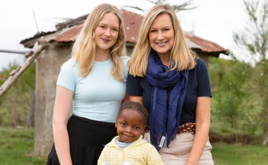 Two women and a child smiling outdoors, with a rustic building and trees in the background.
