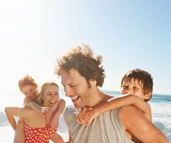 A happy family enjoys a sunny day at the beach, with children on parents' backs, smiling against the ocean backdrop.
