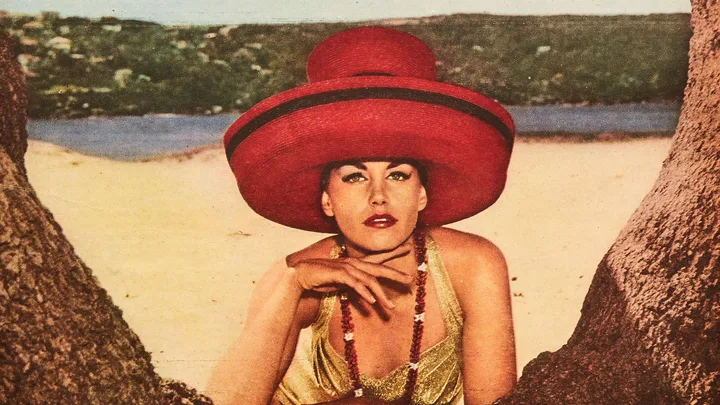 Woman in large red hat and yellow dress, posing between tree trunks on a sandy beach with green hills and water in background.