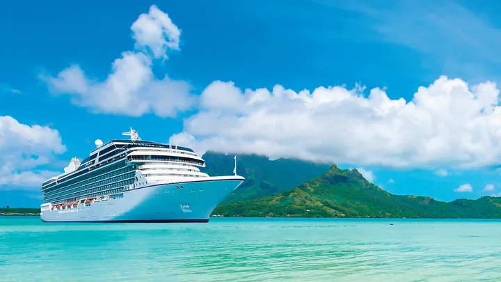 Cruise ship sailing in turquoise waters with a mountainous island and blue sky in the background.