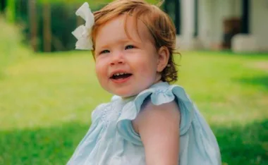 A toddler with red hair and a white bow smiles while sitting on grass, wearing a light blue dress.