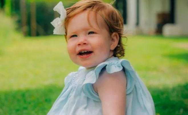 A toddler with red hair and a white bow smiles while sitting on grass, wearing a light blue dress.