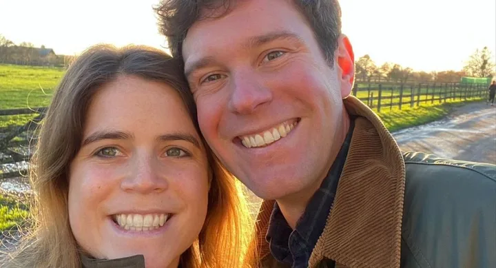 A couple smiling outdoors, with a sunlit field and path in the background.