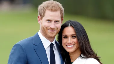 A smiling couple, the man in a blue suit and the woman in a white outfit, standing closely together outdoors.