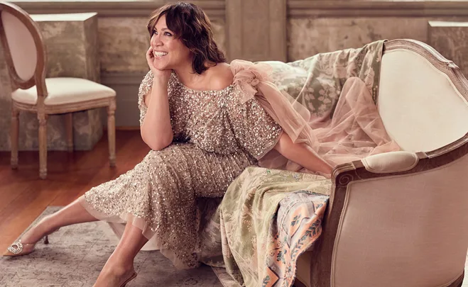 A woman in an elegant sequin dress sits on a vintage sofa, smiling and resting her chin on her hand.