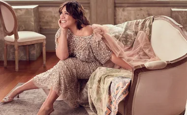 A woman in an elegant sequin dress sits on a vintage sofa, smiling and resting her chin on her hand.