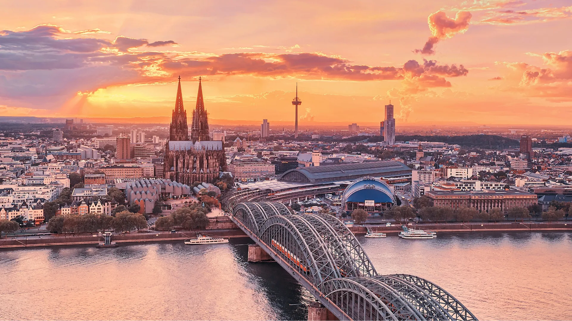 Cologne skyline at sunset, featuring Cologne Cathedral and Hohenzollern Bridge over the Rhine River.