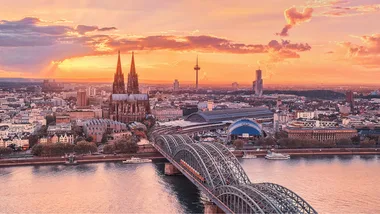 Cologne skyline at sunset, featuring Cologne Cathedral and Hohenzollern Bridge over the Rhine River.