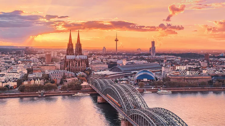 Cologne skyline at sunset, featuring Cologne Cathedral and Hohenzollern Bridge over the Rhine River.