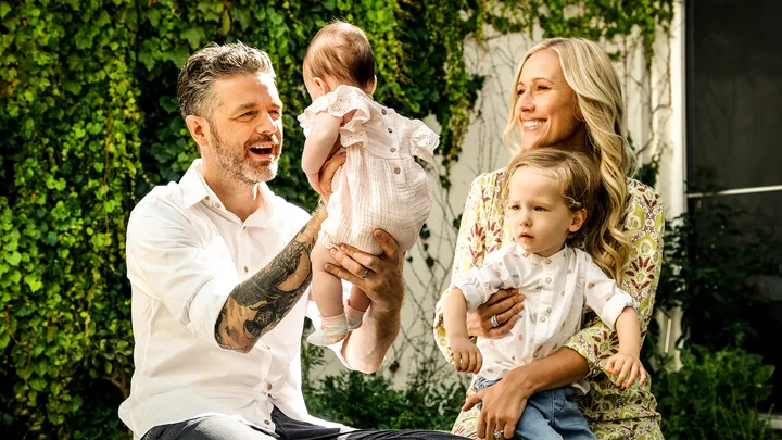 A smiling couple in a garden holds two young children, with green leaves in the background.