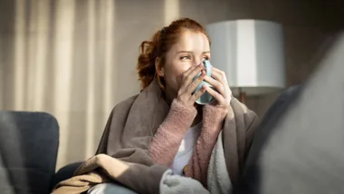 Woman wrapped in blanket holding mug, sitting on couch in cozy room.