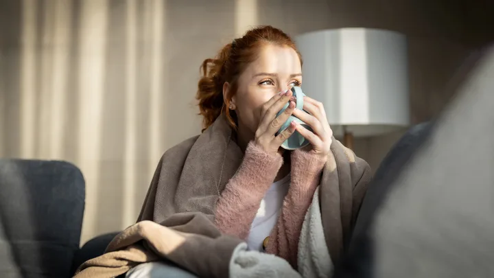 Woman wrapped in blanket holding mug, sitting on couch in cozy room.