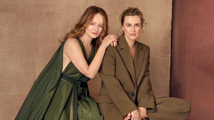 Two women in elegant green and brown outfits pose against a neutral background, with one resting her hand on the other's shoulder.
