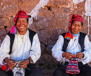 Two men in traditional Peruvian attire knitting, seated against a stone wall, smiling warmly.