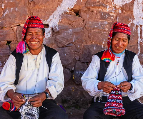 Two men in traditional Peruvian attire knitting, seated against a stone wall, smiling warmly.