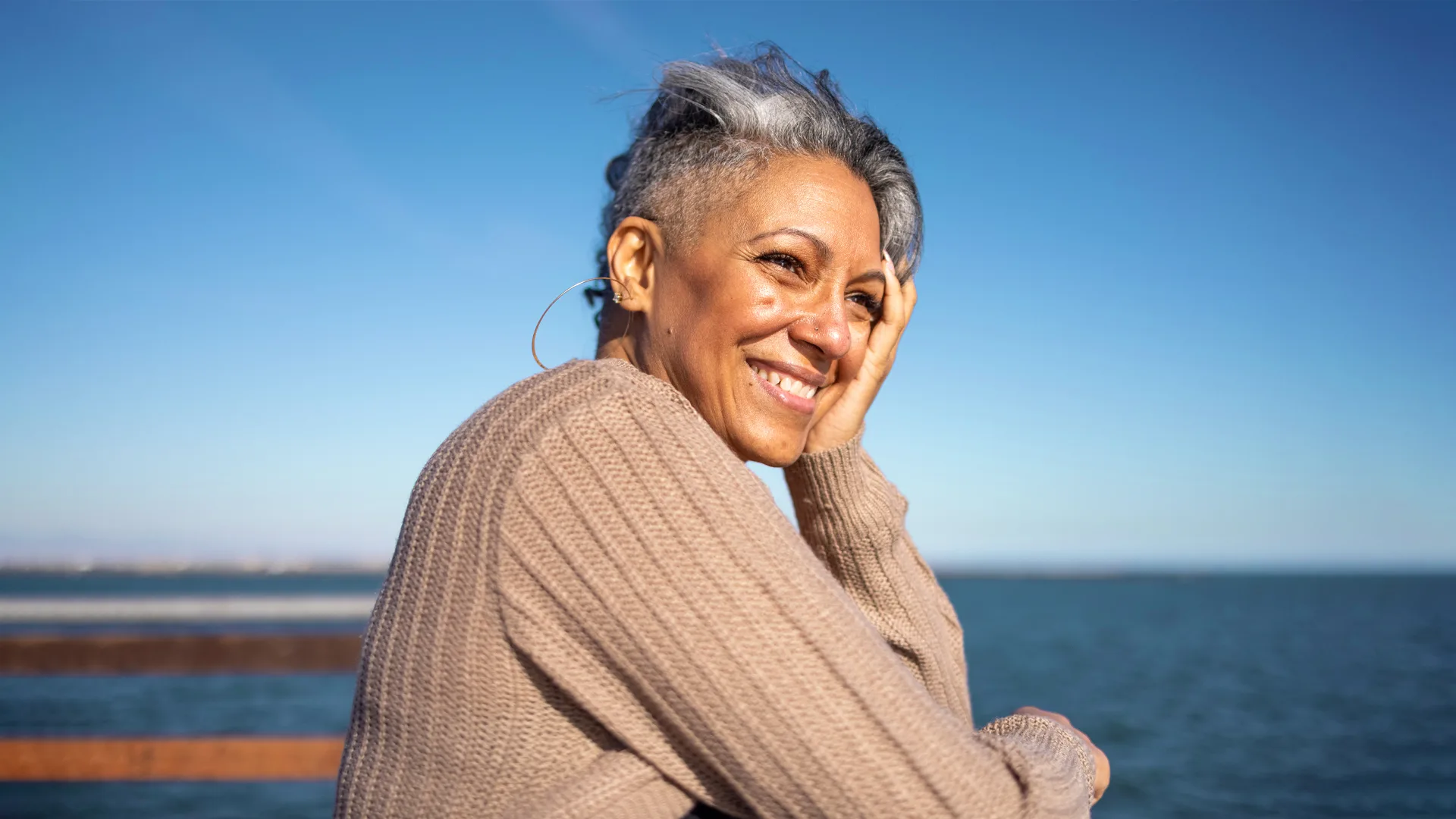 Smiling woman in a sweater with gray hair stands by the water under a clear blue sky.