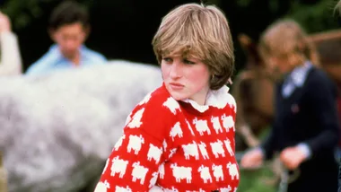 A person wearing a red jumper with white sheep pattern, outdoors with others blurred in the background.