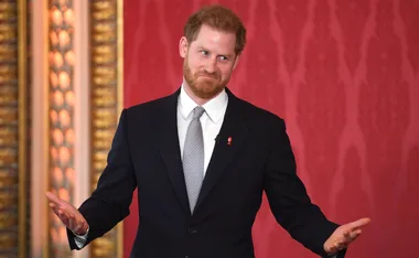 Man in suit with beard gesturing, standing against a red ornate background.