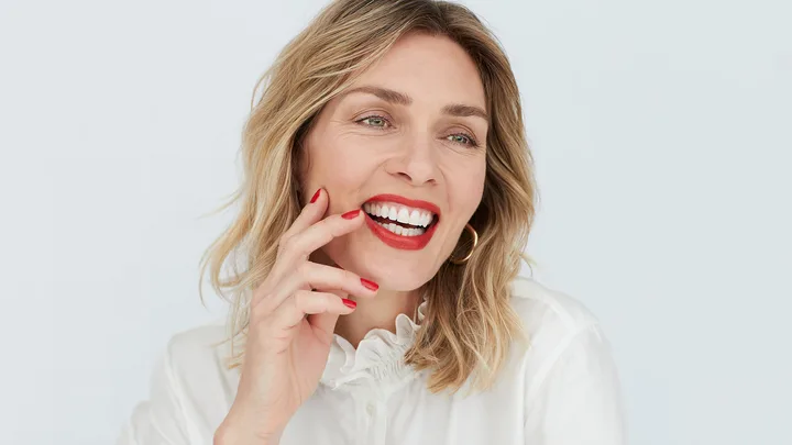 A woman with blond hair smiling, wearing a white shirt and hoop earrings, touching her face with red-painted nails.