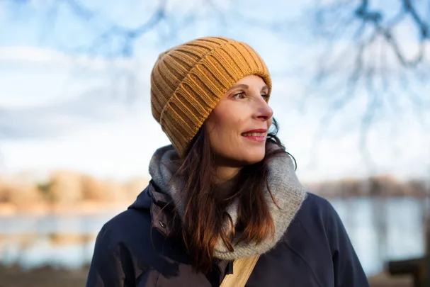 Woman in a yellow beanie and coat, standing outdoors with a blurred background of trees and water.