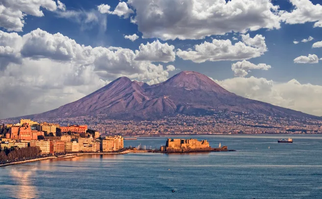 Scenic view of Mount Vesuvius with clouds, overlooking the Gulf of Naples and city buildings on the coastline.