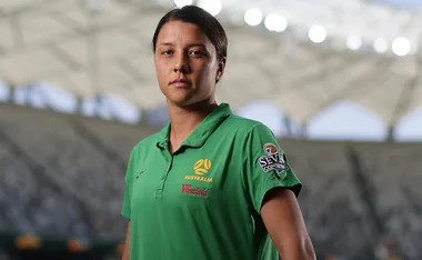 Sam Kerr, wearing an Australian green jersey, stands in a stadium, looking determined.