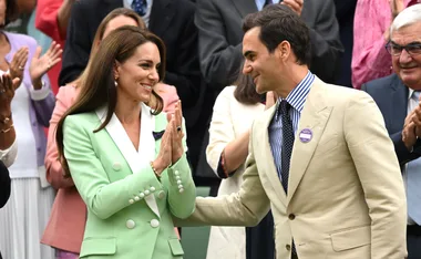 A woman in a green blazer claps while standing with a man in a beige suit, surrounded by applauding crowd.