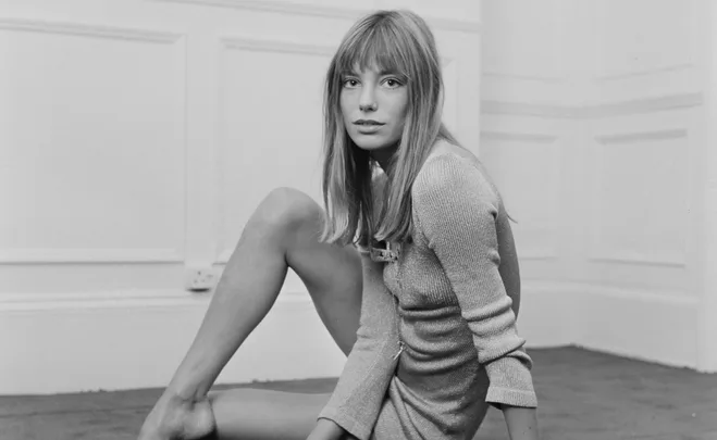 A black and white image of a woman with long hair sitting on the floor, wearing a short dress and looking at the camera.