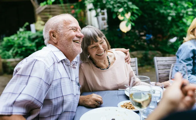 Elderly couple laughing and embracing at an outdoor dining table with wine glasses and plates.