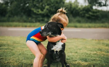 Young child in a colorful swimsuit hugging a black and white dog on a grassy area near a road.
