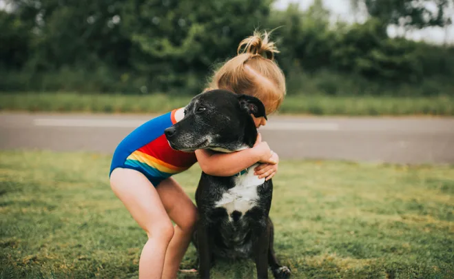 Young child in a colorful swimsuit hugging a black and white dog on a grassy area near a road.