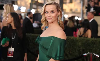 Reese Witherspoon in a green off-shoulder dress at a red carpet event, smiling with people in the background.