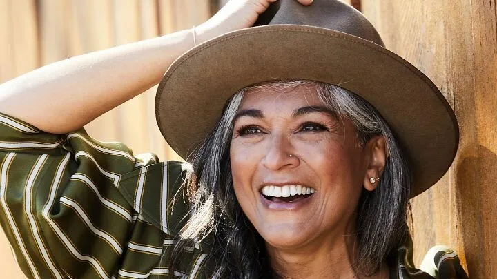An older woman smiles brightly, wearing a striped shirt and a wide-brimmed hat, standing against a wooden background.